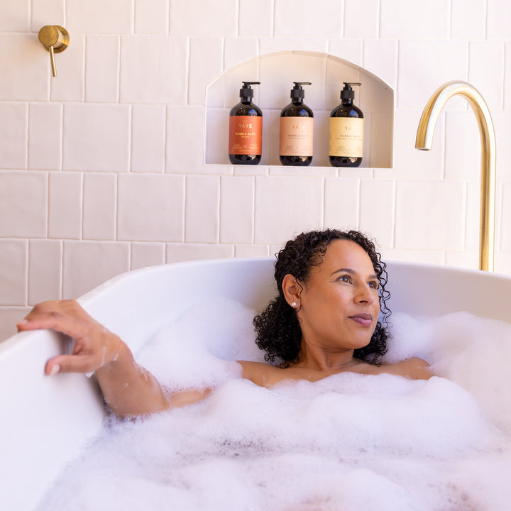 Woman relaxing in a bubble bath with bath products on a shelf above.