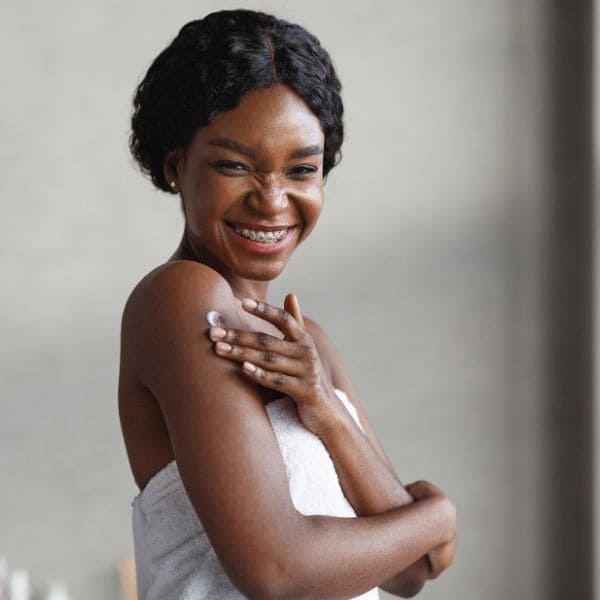 A smiling woman wrapped in a white towel applies moisturiser to her shoulder, showcasing healthy, glowing skin and a joyful self-care moment.